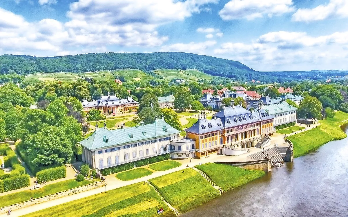 Luftaufnahme des Schlosses Pillnitz an einem sonnigen Tag mit Bergen im Hintergrund.