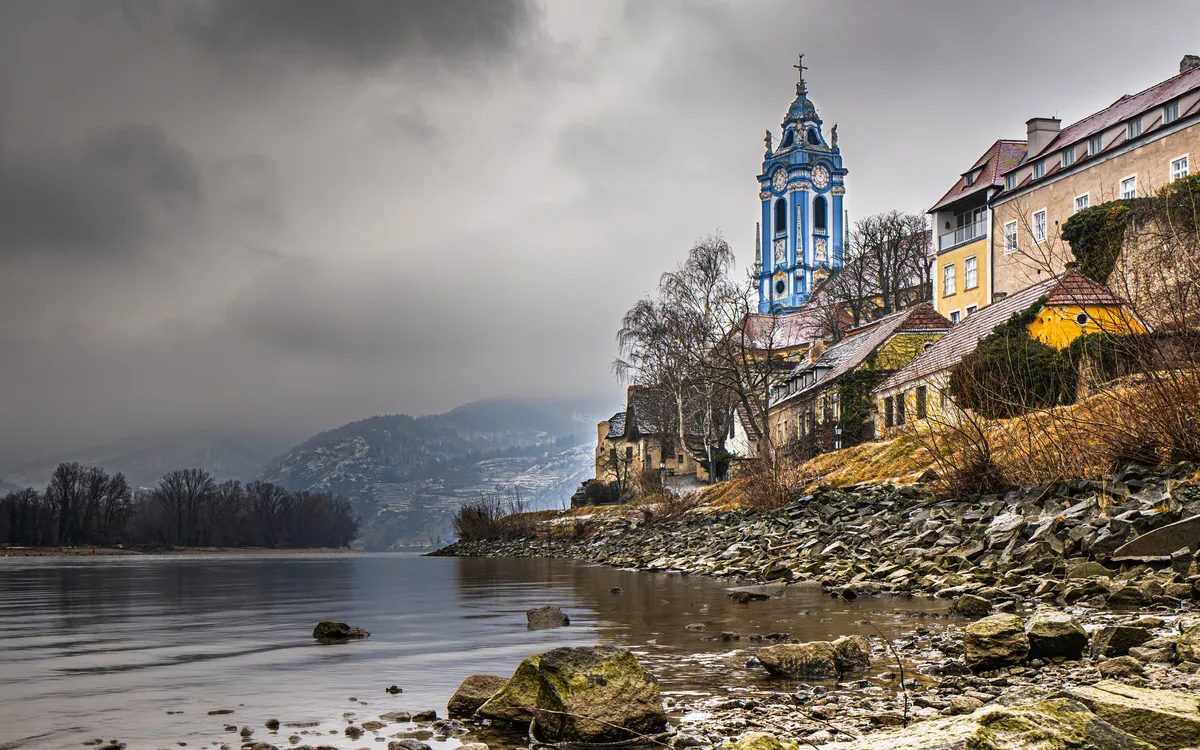 Kirche an einem Flussufer mit bewölktem Himmel und Hügeln im Hintergrund.