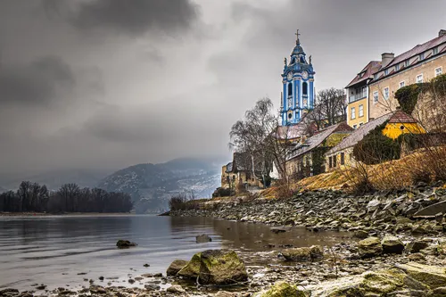 Kirche an einem Flussufer mit bewölktem Himmel und Hügeln im Hintergrund.
