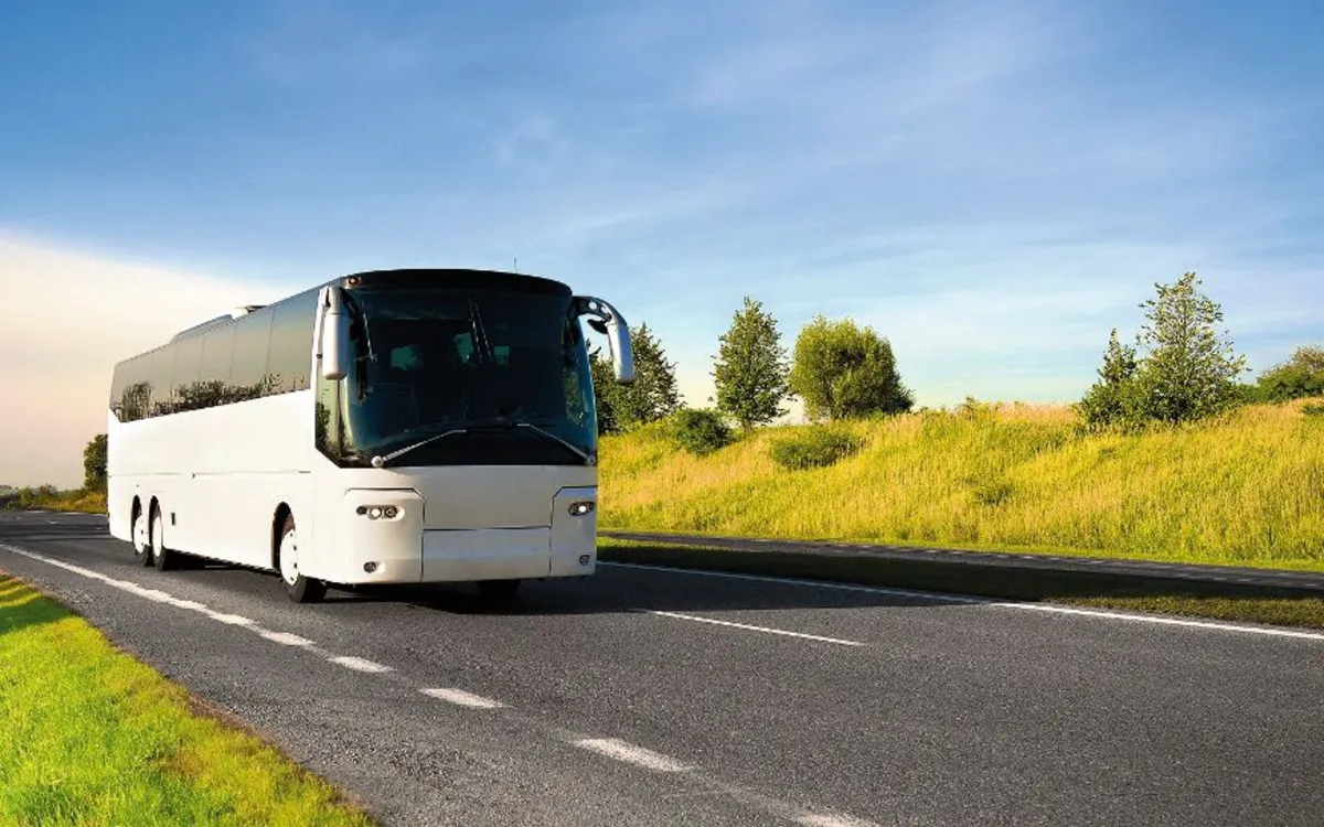 Reisebus fährt auf einer Landstraße bei sonnigem Wetter, umgeben von grünen Wiesen und Bäumen.