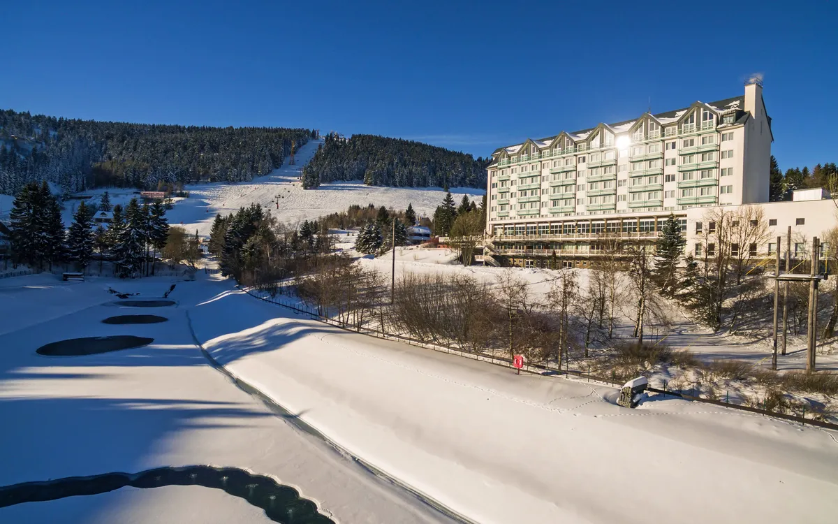 Hotel im Winter mit schneebedecktem Gelände und bewaldeten Hügeln im Hintergrund.