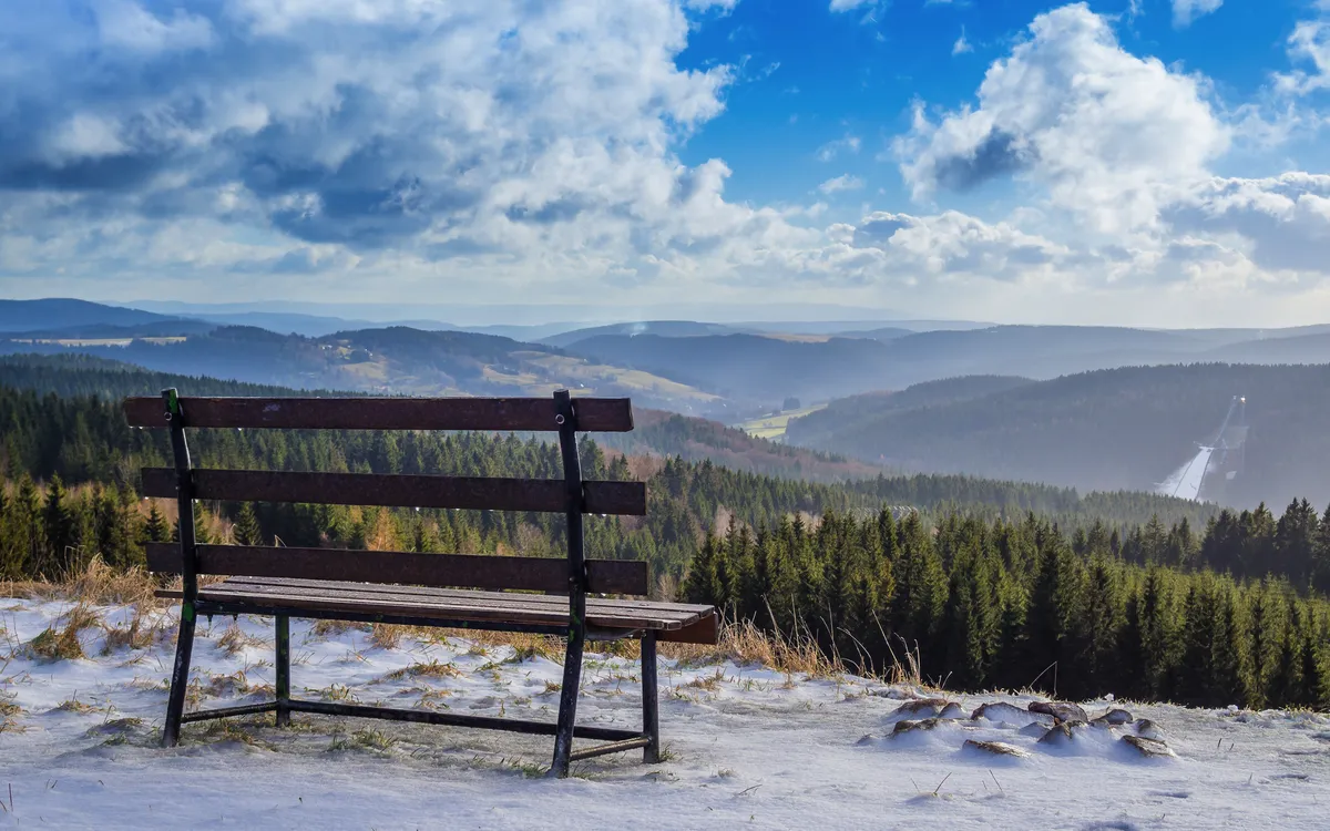 Bank auf einem schneebedeckten Hügel mit Blick auf bewaldete Berge und bewölkten Himmel.