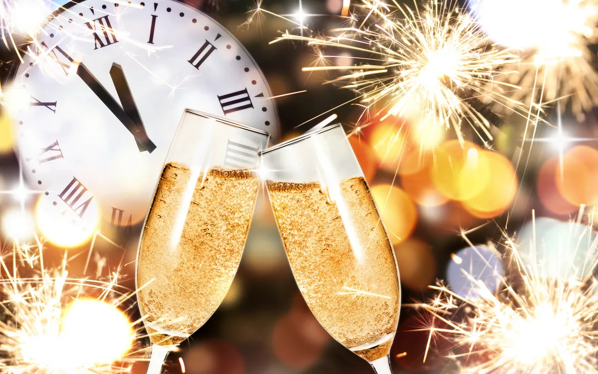 Zwei Sektgläser stoßen an, umgeben von Feuerwerk und Uhr zeigt Mitternacht.