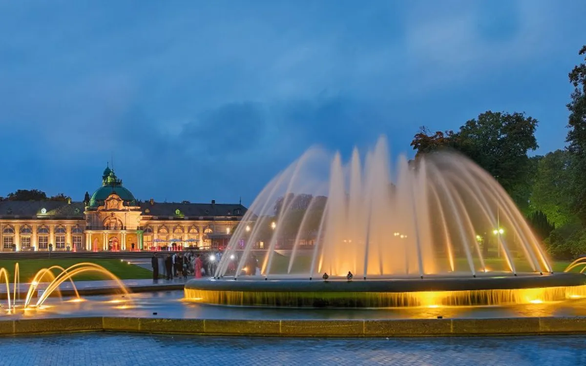 Springbrunnen in einem Park bei Dämmerung mit beleuchtetem Gebäude im Hintergrund.