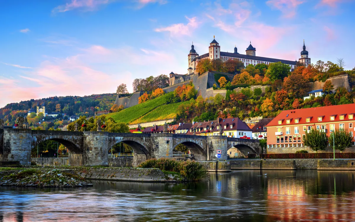Festung auf einem Hügel mit Brücke im Vordergrund, herbstliche Bäume im Hintergrund.