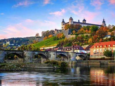 Festung auf einem Hügel mit Brücke im Vordergrund, herbstliche Bäume im Hintergrund.