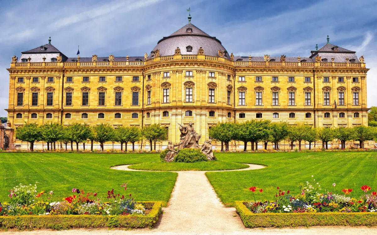 Barockes Schloss mit gepflegtem Garten und blühenden Blumenbeeten im Vordergrund.