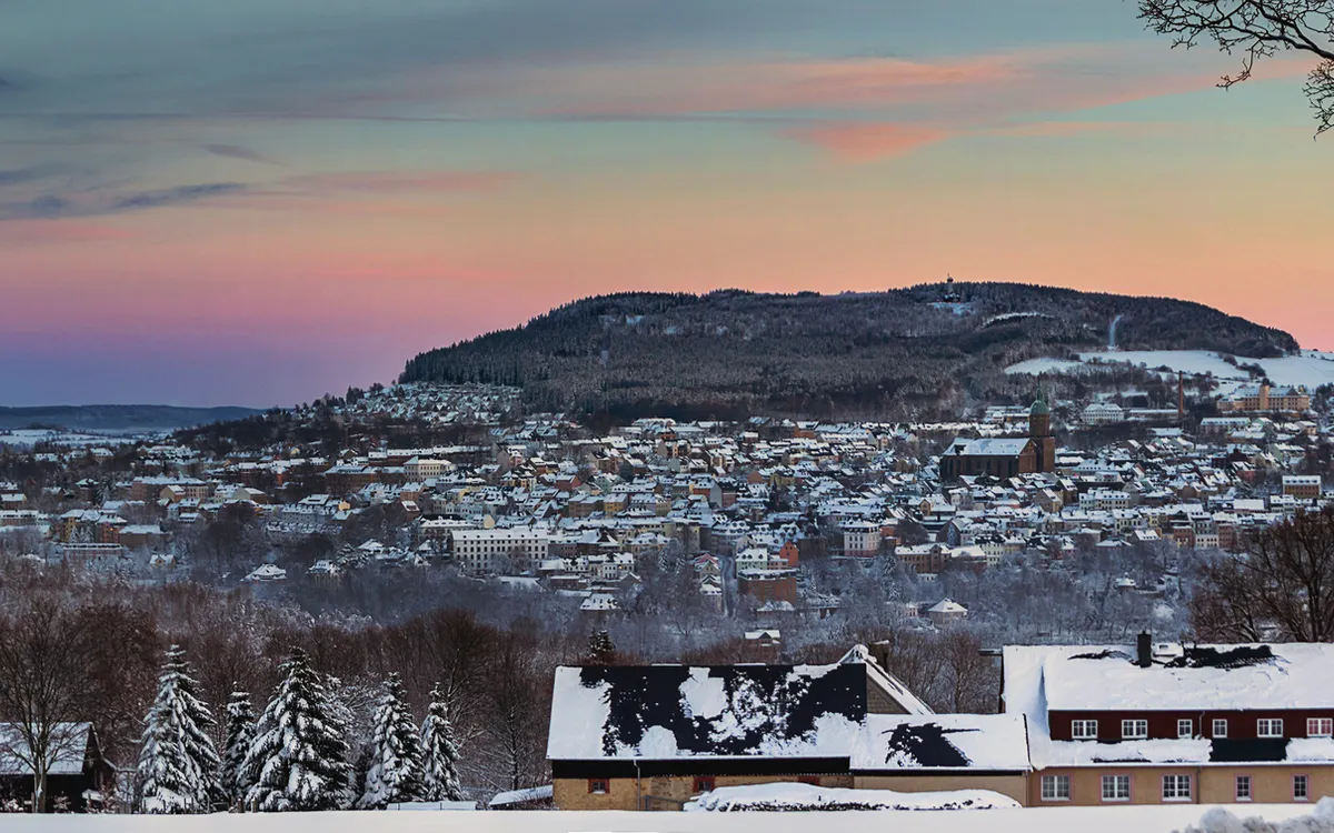 Winterliche Stadtansicht bei Sonnenuntergang mit schneebedeckten Häusern und Hügel im Hintergrund.
