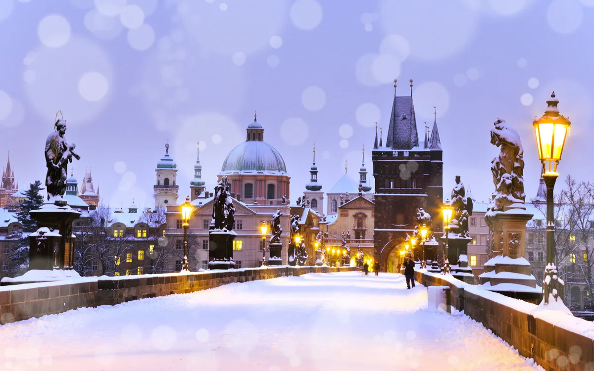 Verschneite Karlsbrücke in Prag mit erleuchteten Laternen und historischer Kulisse.