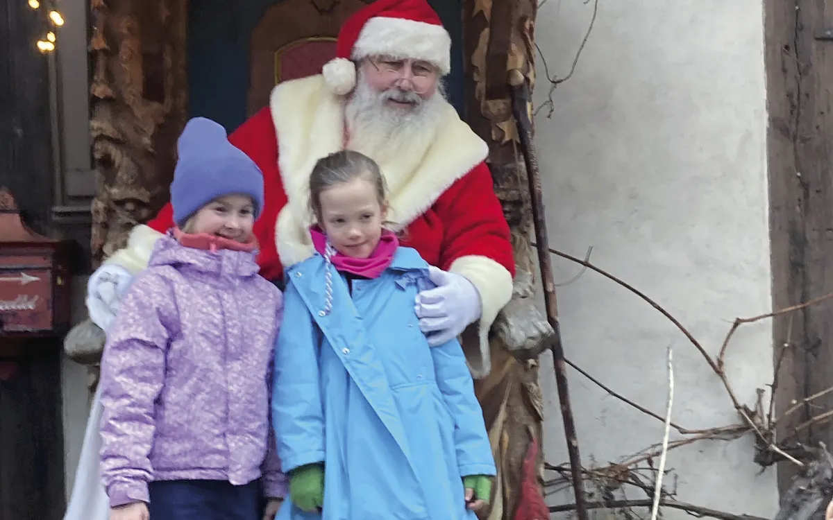 Zwei Kinder posieren mit einem Weihnachtsmann vor einer weihnachtlich dekorierten Kulisse.