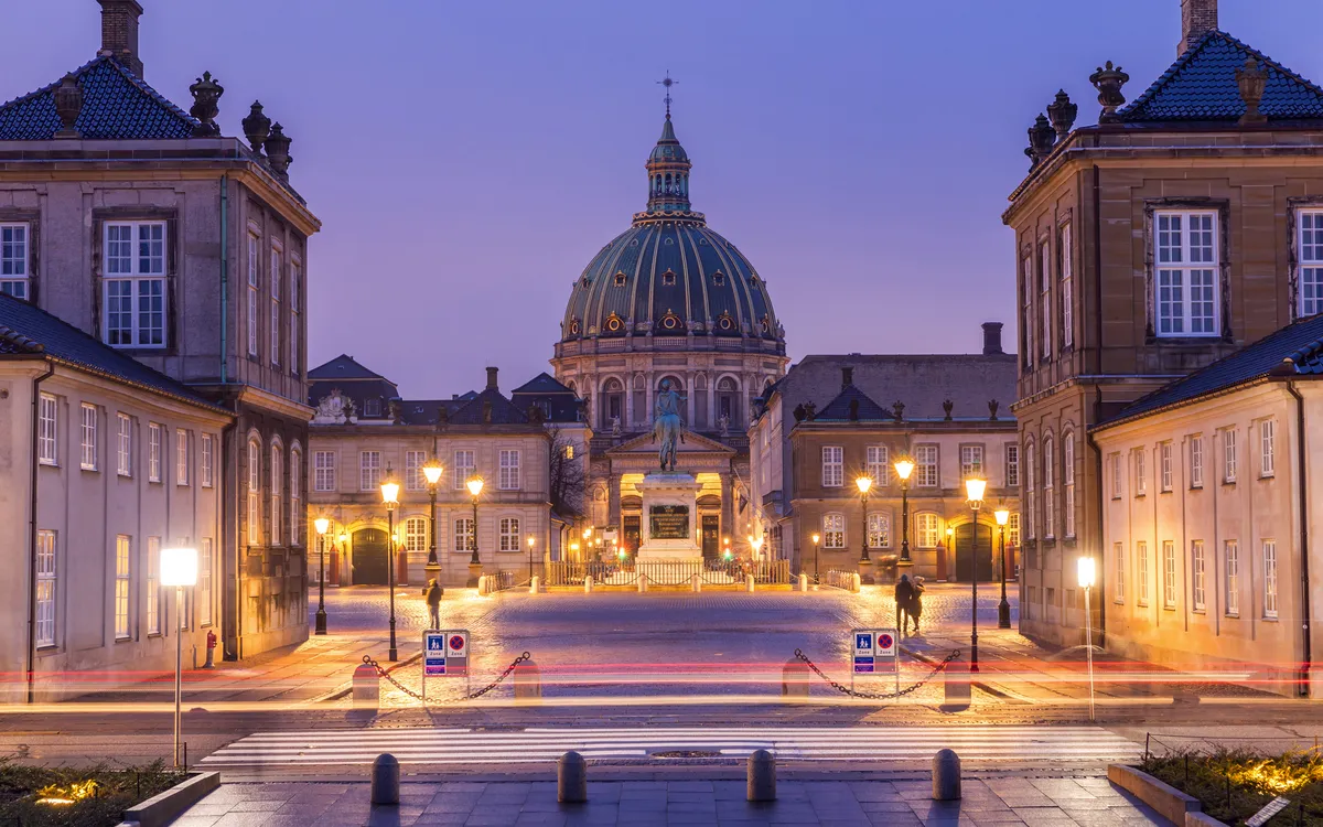 Schloss Amalienborg in Kopenhagen bei Nacht, beleuchtet, mit Wasser im Vordergrund.