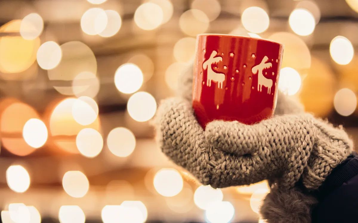 Hand in Wollhandschuhen hält Tasse mit weihnachtlichem Motiv, Lichter im Hintergrund.
