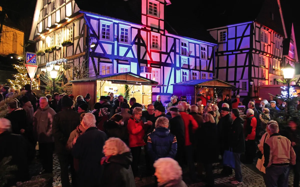 Menschenmenge bei Nacht vor beleuchteten Fachwerkhäusern auf einem Weihnachtsmarkt.