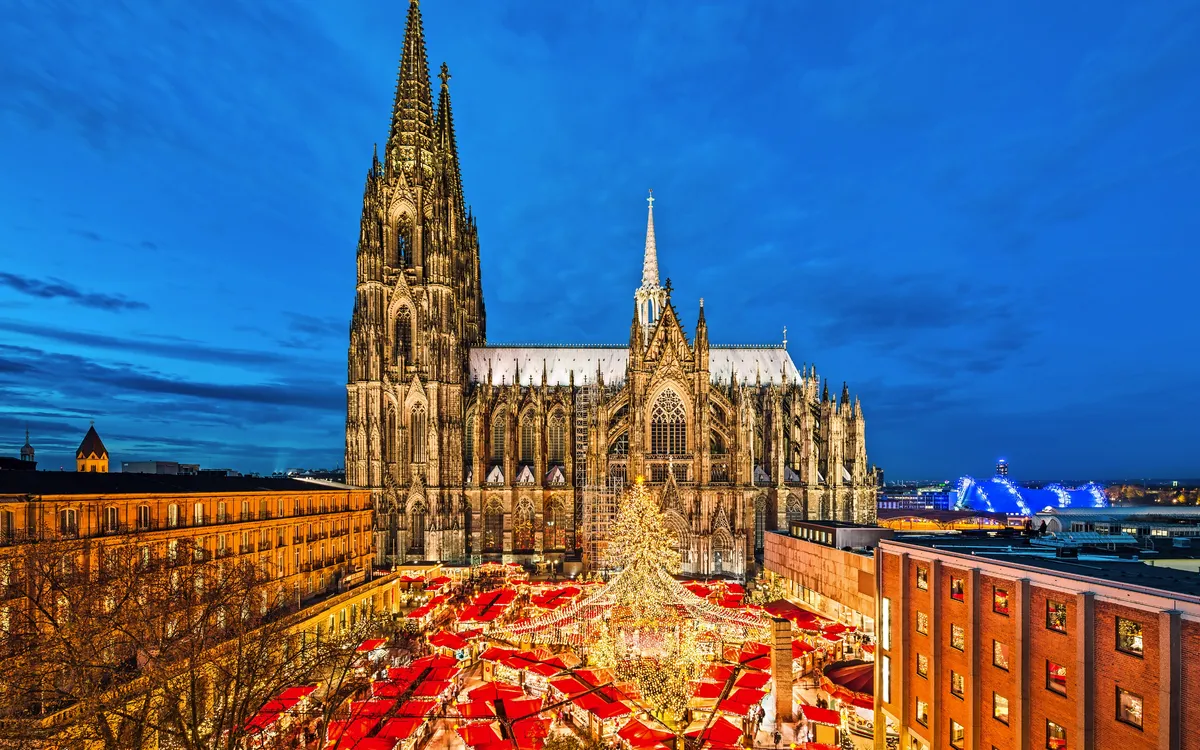 Kölner Dom bei Nacht, beleuchteter Weihnachtsmarkt im Vordergrund.