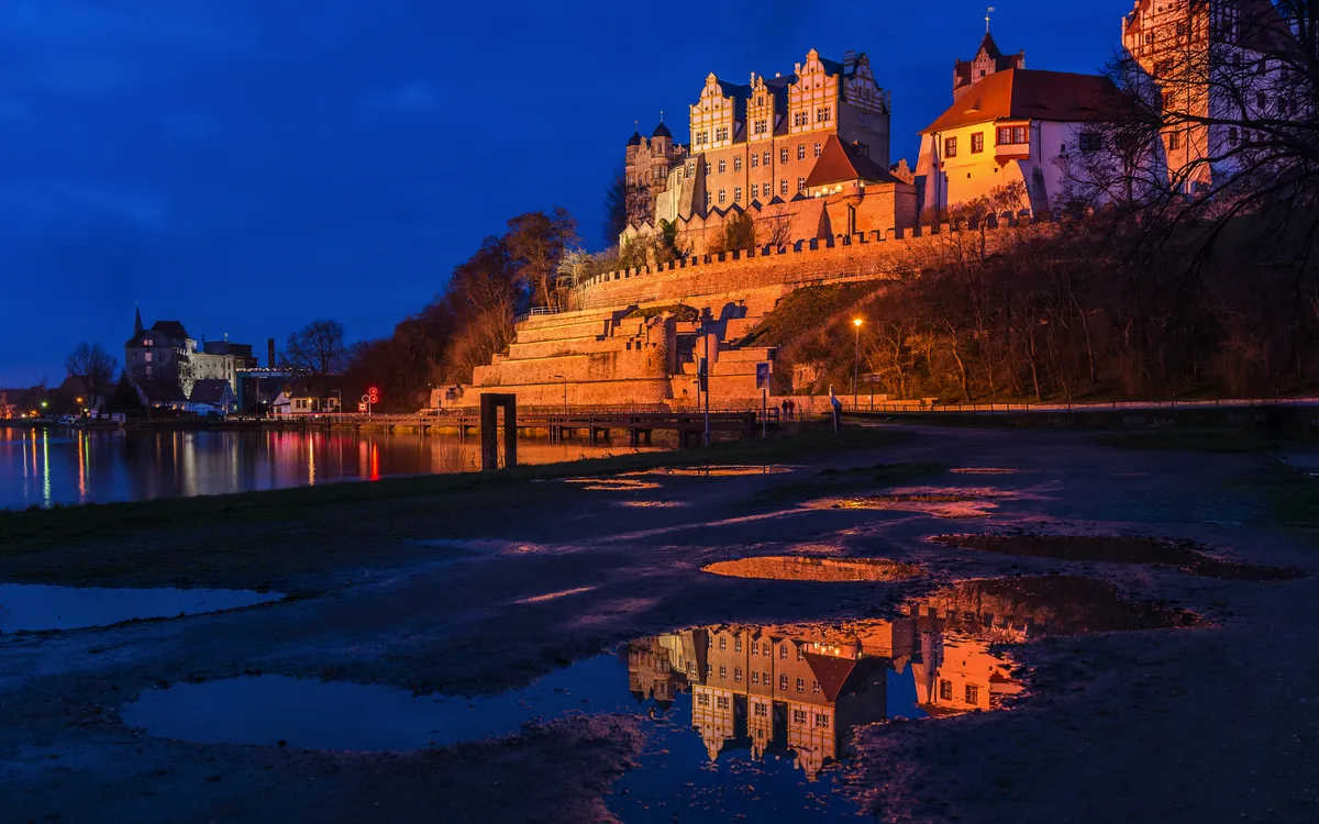 Burg bei Nacht mit Spiegelung im Wasser, beleuchtet von warmem Licht vor blauem Himmel.