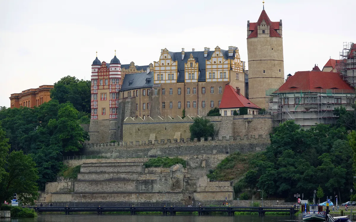 Ein Schloss auf einem Hügel mit umliegenden Bäumen und einem Fluss im Vordergrund.