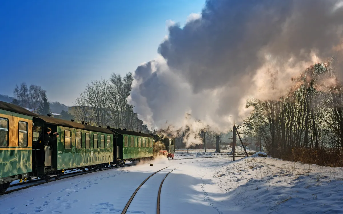 Grüner Dampflokzug auf verschneiter Strecke, Rauch steigt empor, Bäume im Hintergrund.