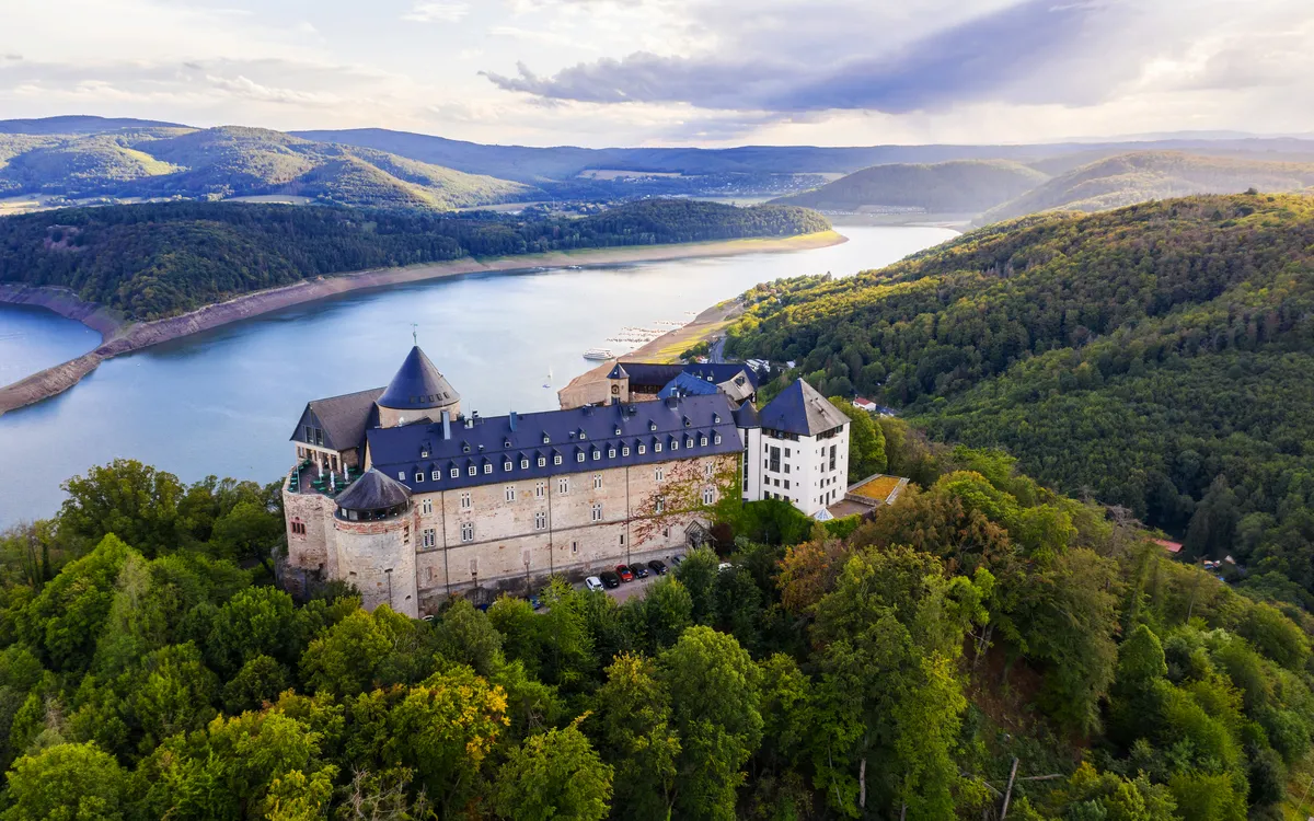 Luftaufnahme einer historischen Burg umgeben von Wald und See in hügeliger Landschaft