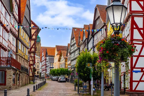 Gepflasterte Straße mit Fachwerkhäusern und Blumen an einer Laterne.