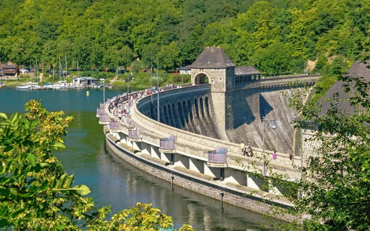 Staumauer mit Menschen, grünem Wald und Wasser im Hintergrund