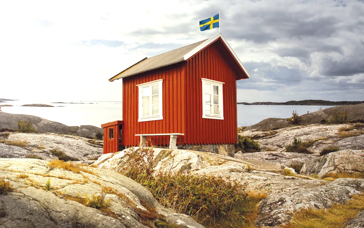 Rotes schwedisches Holzhaus am Meer mit schwedischer Flagge, Felsenlandschaft im Hintergrund