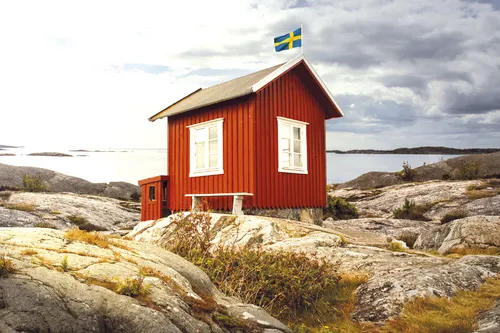 Rotes schwedisches Holzhaus am Meer mit schwedischer Flagge, Felsenlandschaft im Hintergrund