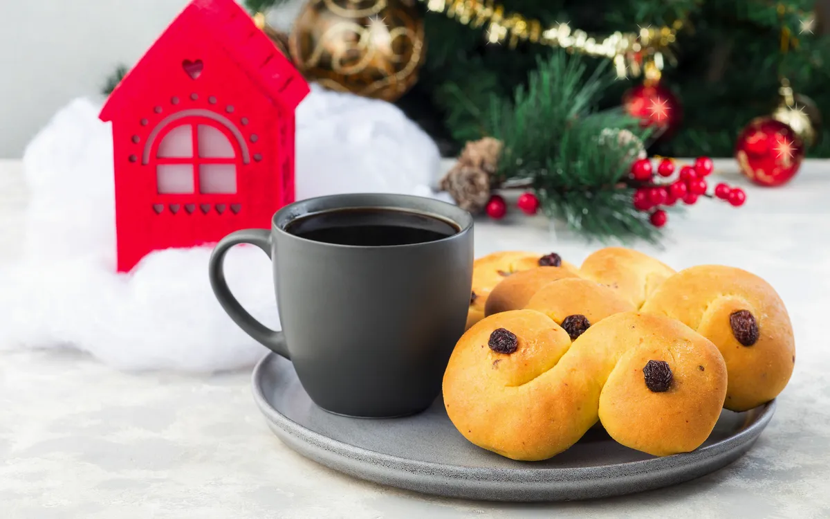 Lussekatter und Tasse Kaffee vor weihnachtlicher Dekoration.