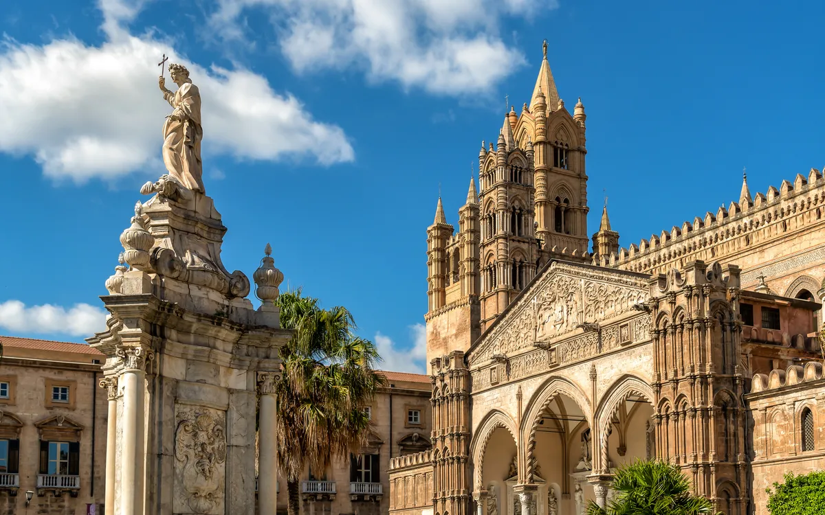 Statue und Kathedrale mit verzierten Fassaden und blauem Himmel im Hintergrund.