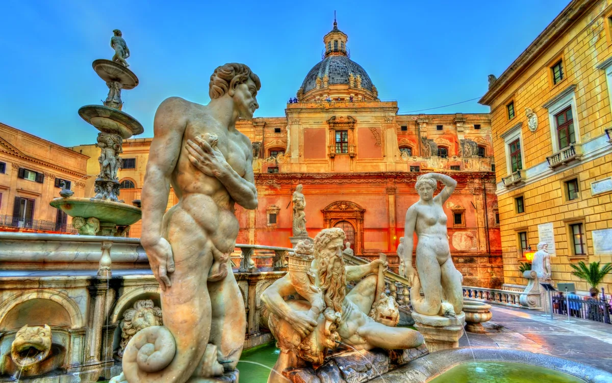 Barocker Springbrunnen mit Skulpturen vor historischem Gebäude bei Sonnenuntergang.