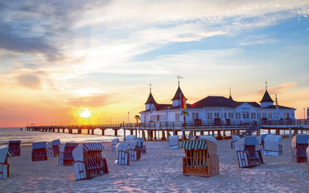 Strandkörbe am Sandstrand vor Seebrücke bei Sonnenuntergang mit pastellfarbenem Himmel.