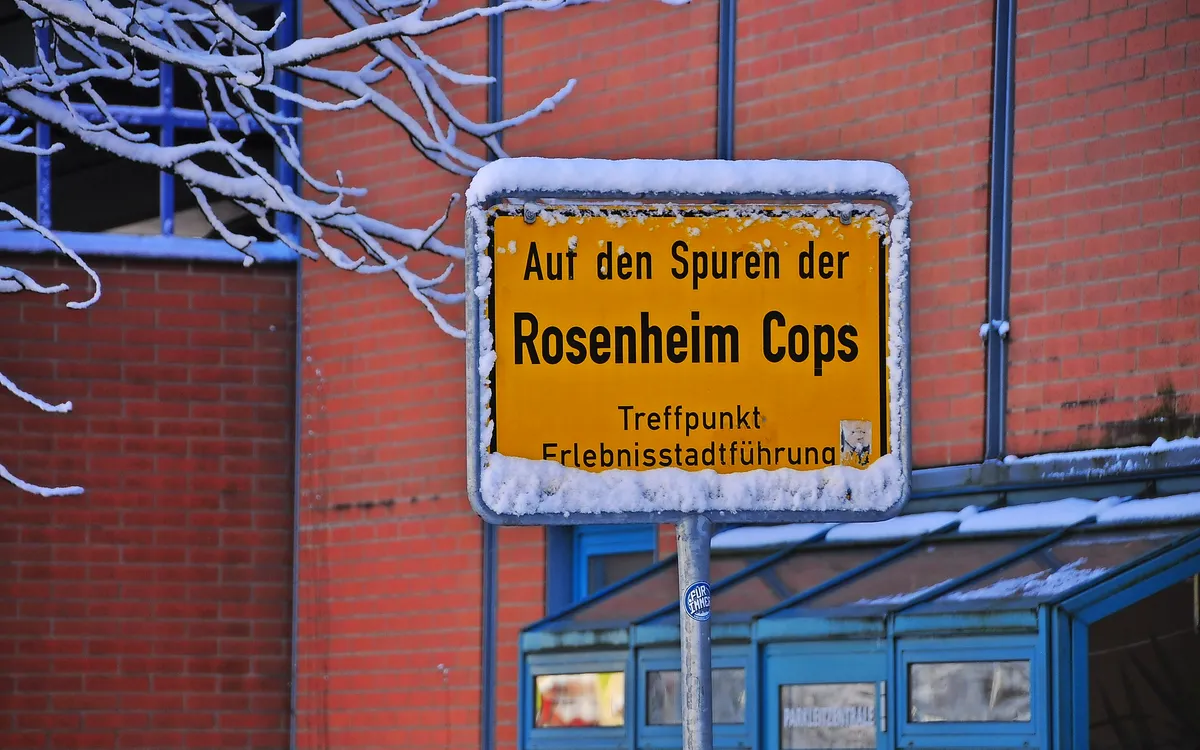 Schneebedecktes Schild mit Aufschrift "Auf den Spuren der Rosenheim Cops" vor Backsteinwand.
