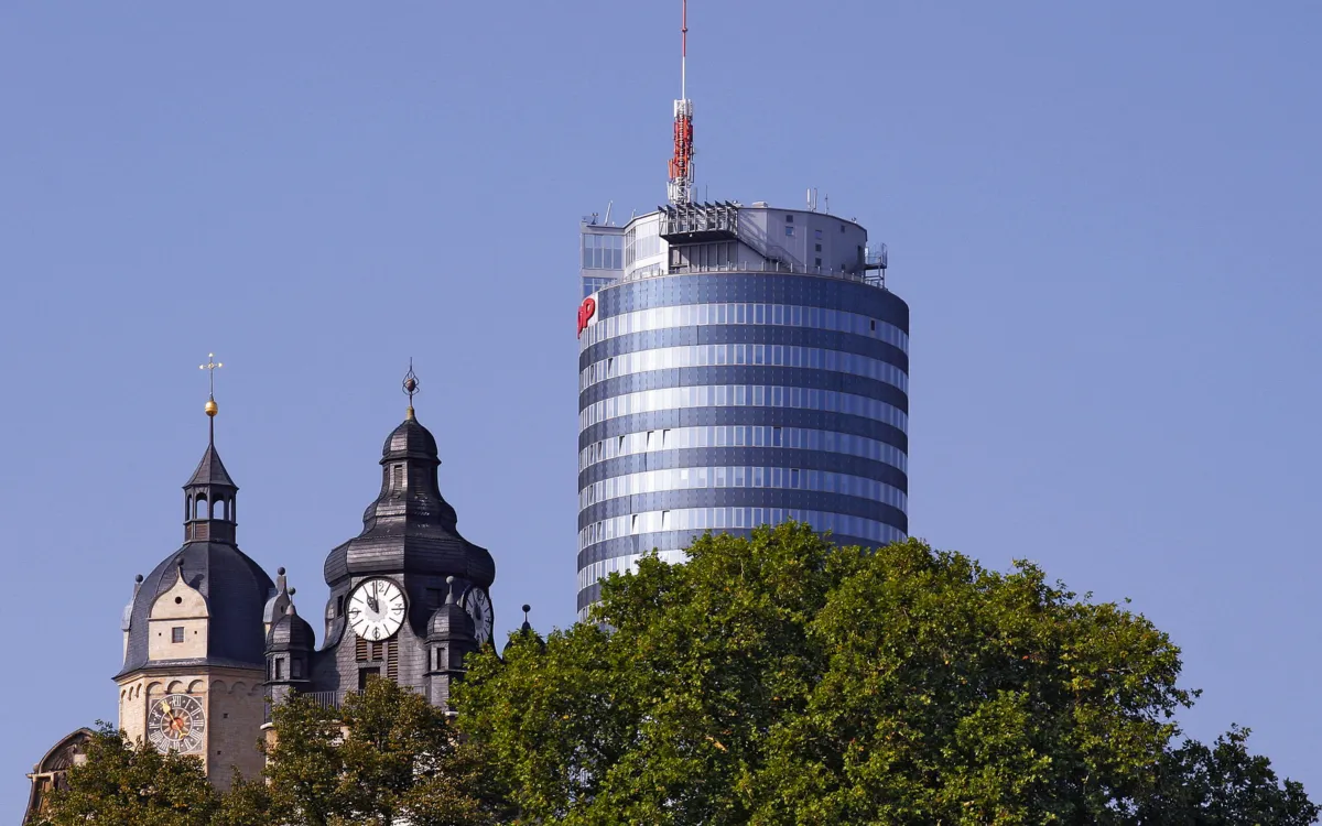 Kirchturm und modernes Gebäude hinter Baumkronen bei blauem Himmel.
