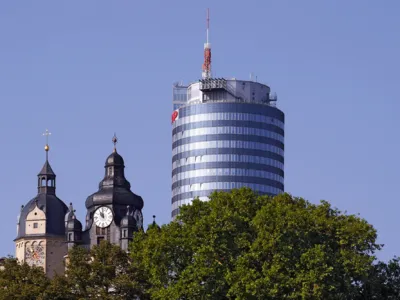 Kirchturm und modernes Gebäude hinter Baumkronen bei blauem Himmel.