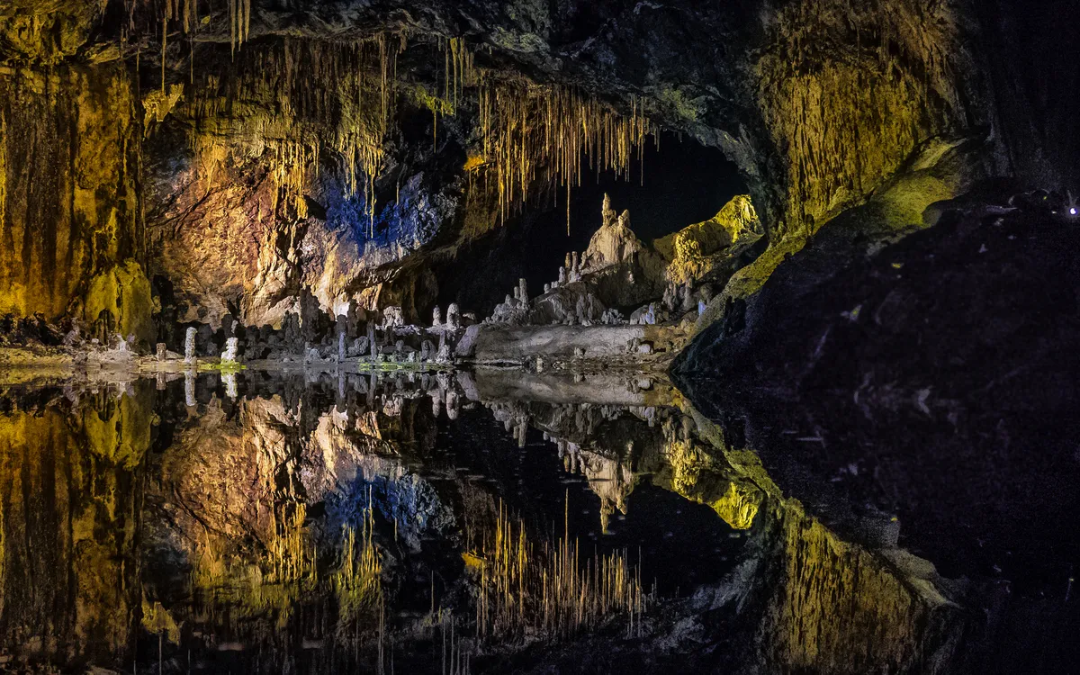 Spiegelung einer beleuchteten Höhlenwand mit Stalagmiten im Wasser.