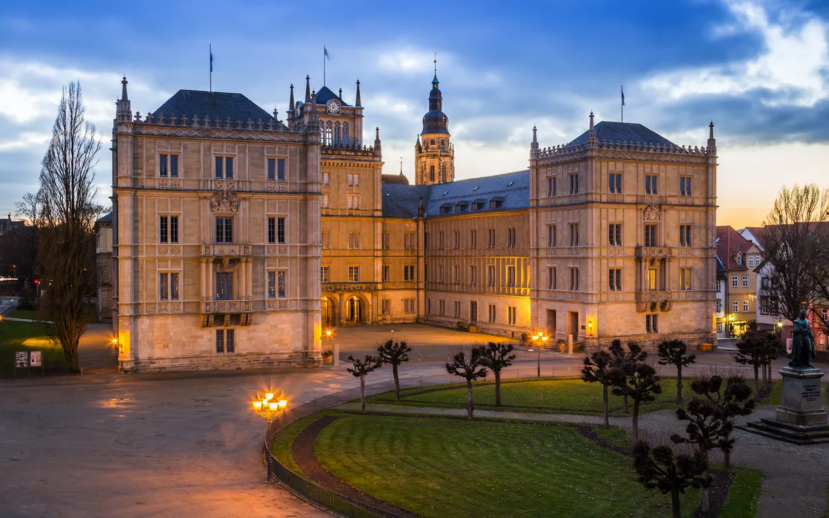 Historisches Schlossgebäude bei Dämmerung mit beleuchteten Fenstern und wolkigem Himmel.