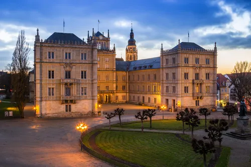 Historisches Schlossgebäude bei Dämmerung mit beleuchteten Fenstern und wolkigem Himmel.