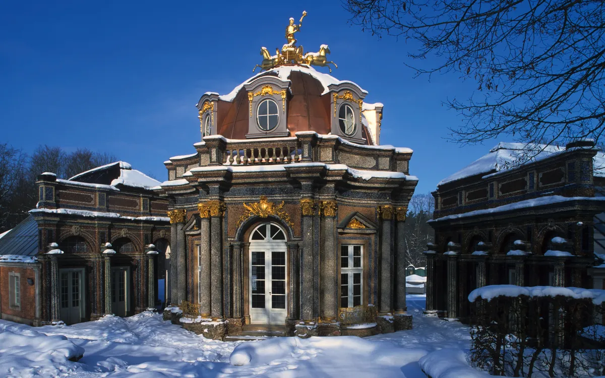 Barockpavillon im Schnee mit blauer Himmel und goldener Statue.