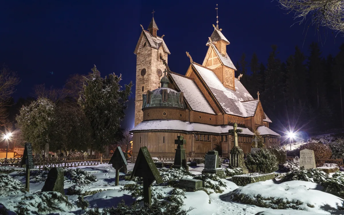Schnee bedeckte Holzkirche bei Nacht mit Grabsteinen im Vordergrund.