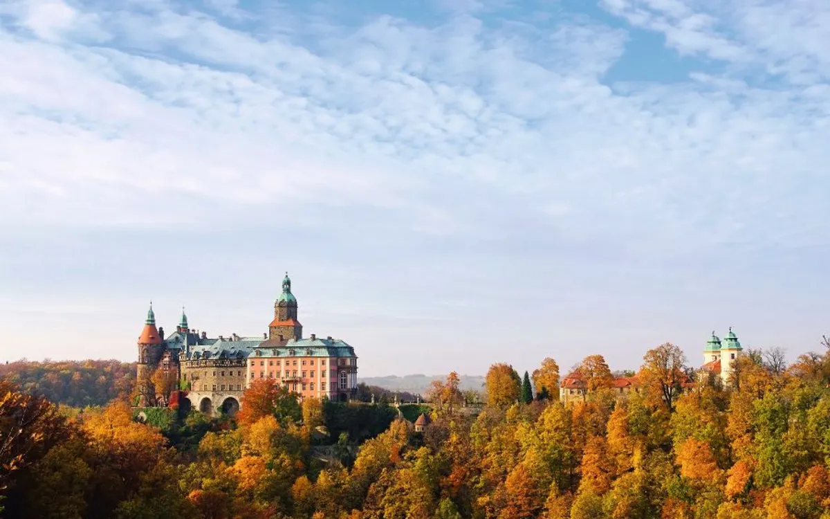 Schloss auf einem Hügel, umgeben von herbstlichem Wald, unter teils bewölktem Himmel.