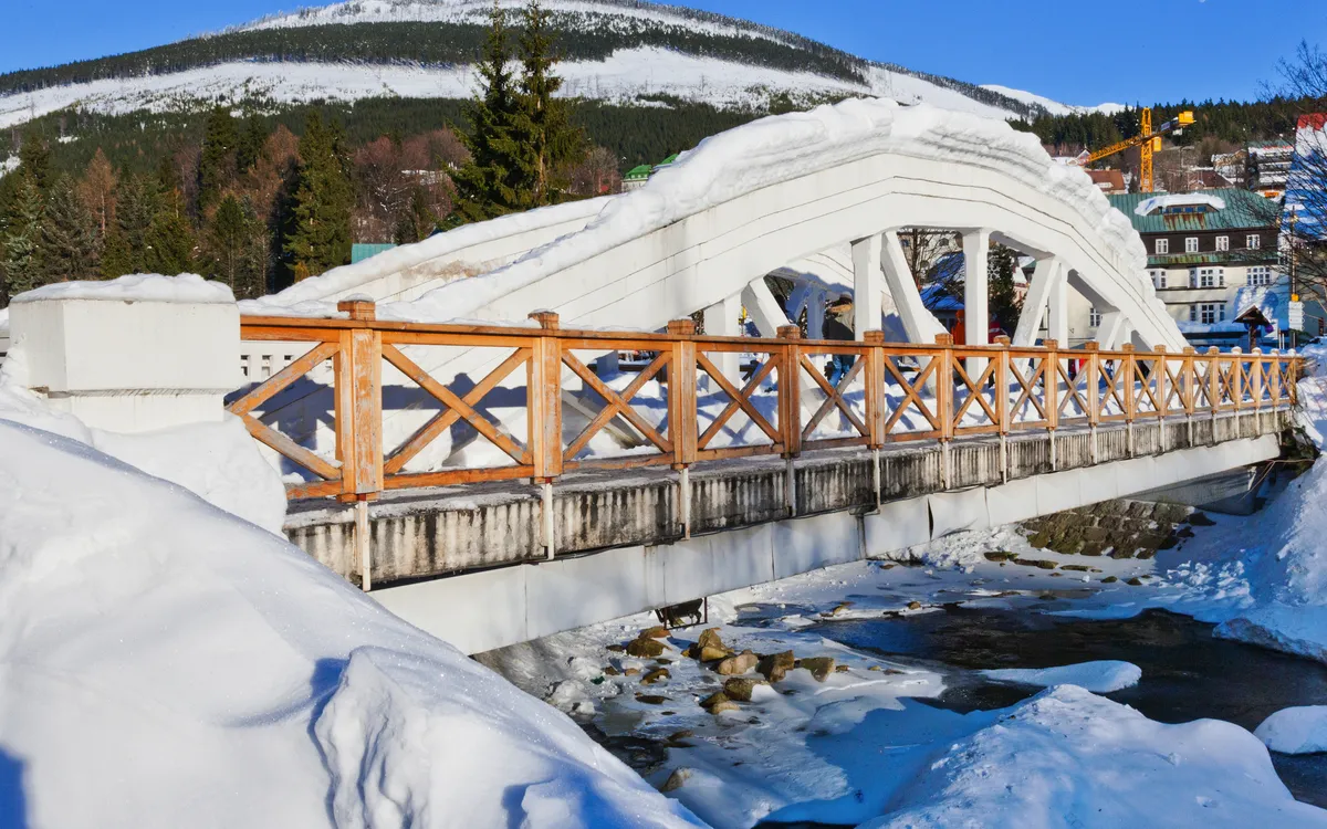 Schnee bedeckte Brücke vor einem bewaldeten, verschneiten Gebirgshügel im Winter.