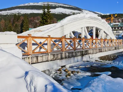 Schnee bedeckte Brücke vor einem bewaldeten, verschneiten Gebirgshügel im Winter.