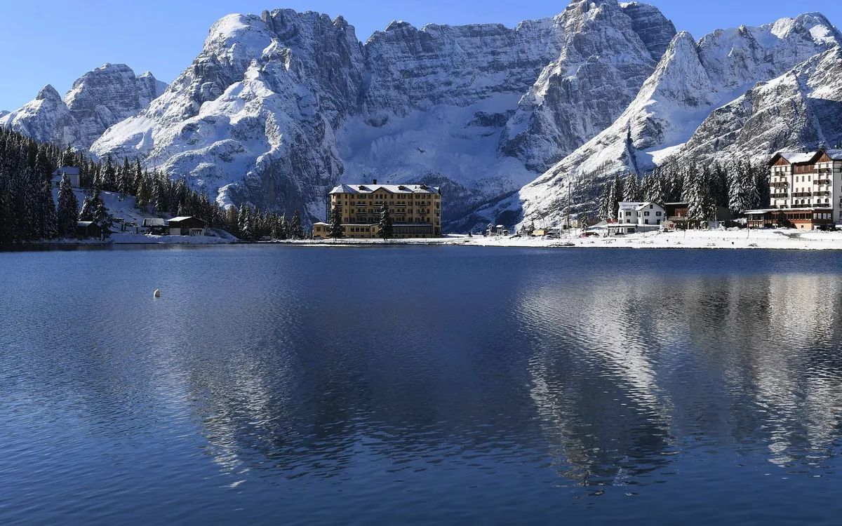 Bergsee mit schneebedeckten Bergen und Gebäuden im Hintergrund.