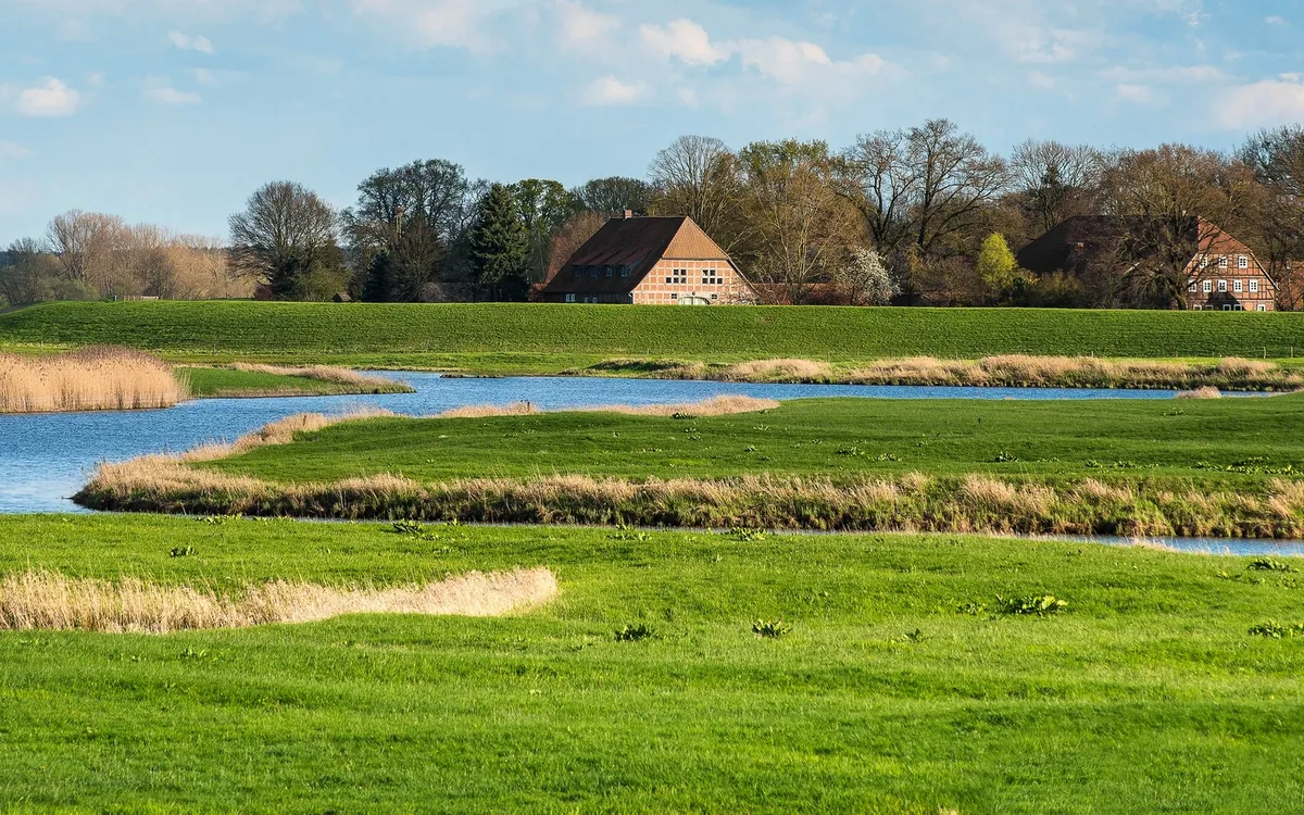 Grüne Wiesen mit Fluss und Bauernhäusern im Hintergrund.