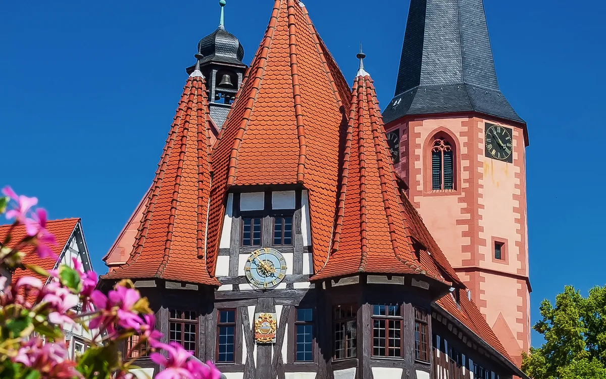 Historisches Fachwerkhaus mit Kirchturm bei blauem Himmel und Blumen im Vordergrund.