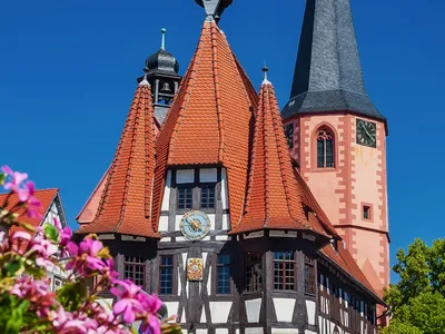 Historisches Fachwerkhaus mit Kirchturm bei blauem Himmel und Blumen im Vordergrund.