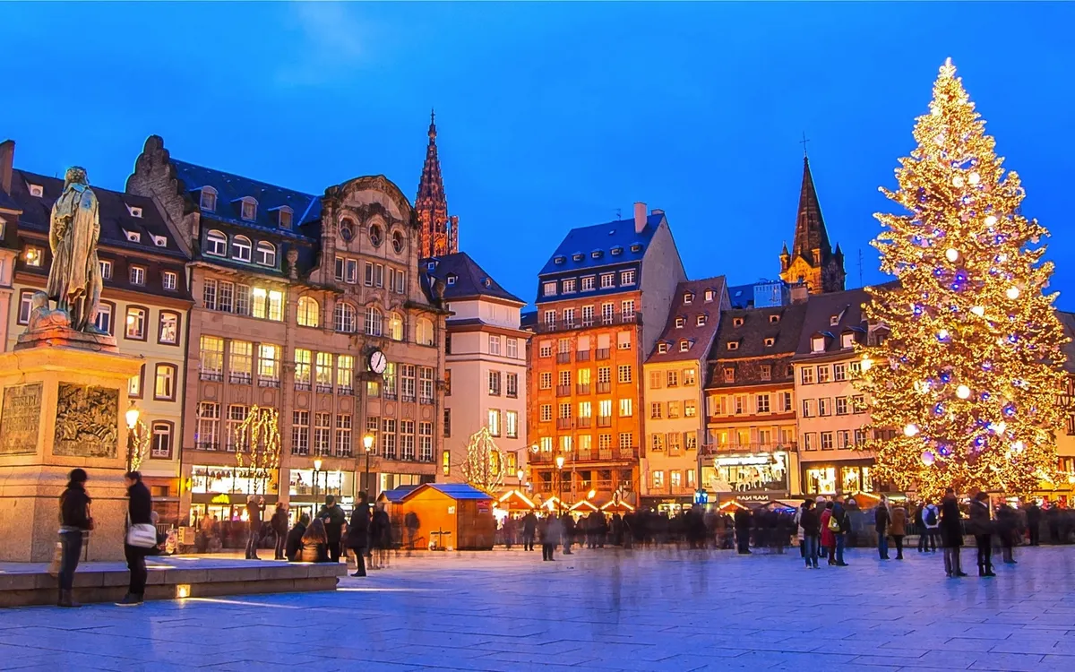 Weihnachtsmarkt auf einem Platz mit großen Christbaum und historischen Gebäuden im Hintergrund.