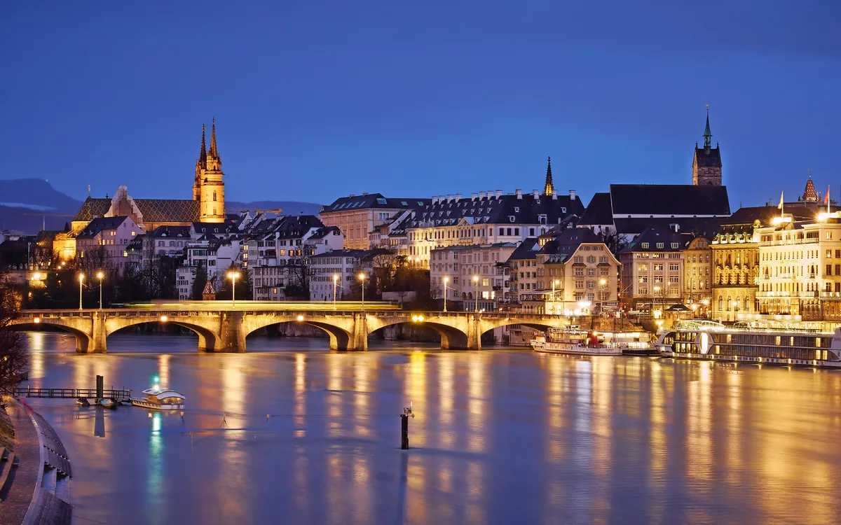Flussansicht der Stadt Basel bei Nacht mit beleuchteten Gebäuden und Brücke.