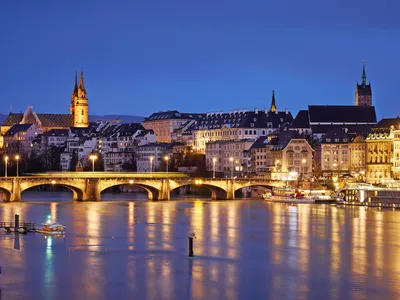 Flussansicht der Stadt Basel bei Nacht mit beleuchteten Gebäuden und Brücke.