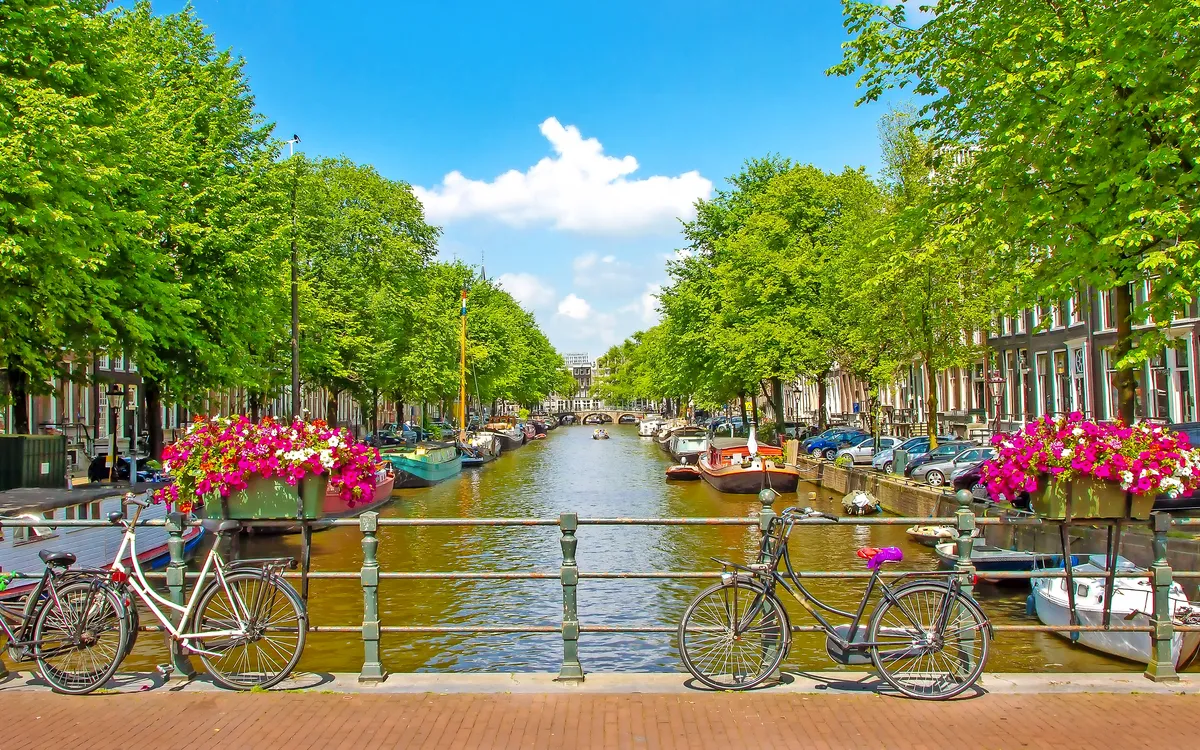 Kanal in Amsterdam mit Blumen, Fahrrädern und Gebäuden bei blauem Himmel.