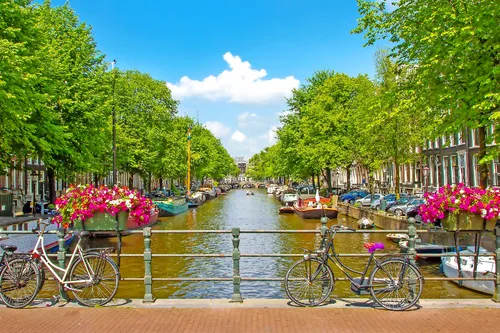 Kanal in Amsterdam mit Blumen, Fahrrädern und Gebäuden bei blauem Himmel.